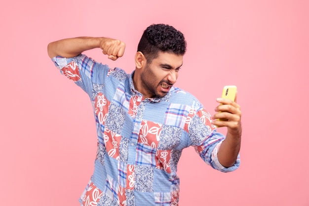 Portrait d'un homme fou en colère avec barbe portant une chemise de style décontracté bleu ayant un appel vidéo désagréable poing fermé pour frapper l'expression folle du téléphone portable Chemise de studio intérieur isolée sur fond rose