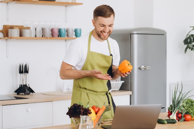 Portrait d'un homme heureux à la maison homme cuisinant une salade de légumes en regardant la caméra et souriant en train de trancher des légumes à l'aide d'un ordinateur portable pour une formation en cuisine en ligne