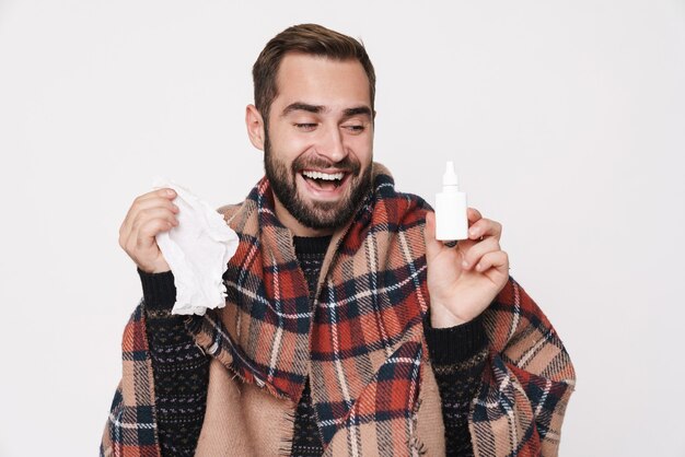 Portrait d'un homme heureux et malade enveloppé dans une couverture éternuant et tenant des gouttes nasales à cause de la grippe isolée sur un mur blanc