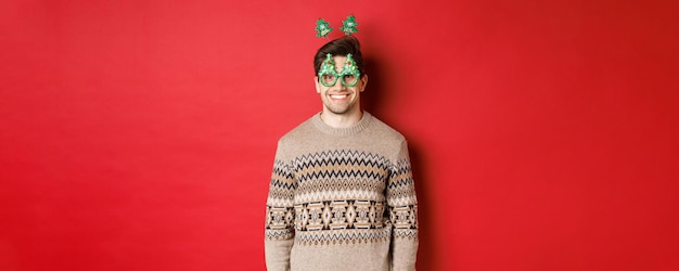 Portrait d'un homme heureux et mignon en pull d'hiver et lunettes de fête, célébrant le nouvel an ou noël, souriant debout sur fond rouge.