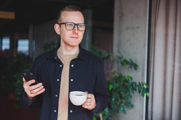 Portrait d'un homme heureux et réussi dans des lunettes optiques tenant un téléphone portable moderne et un café dans ses mains un blogueur joyeux avec un smartphone numérique posant dans un café