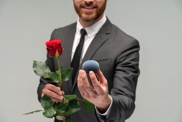 Portrait d'un homme heureux satisfait vêtu d'un costume formel tenant une boîte avec une bague de fiançailles et l'étirant vers la caméra isolée sur fond gris