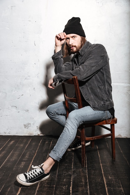 Portrait d'un homme hipster au chapeau assis sur une chaise isolée sur le mur