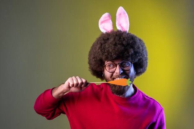 Portrait d'un homme hipster avec une coiffure afro debout mordant une carotte savoureuse et regardant la caméra, portant un sweat-shirt rouge. Prise de vue en studio intérieure isolée sur fond néon coloré.