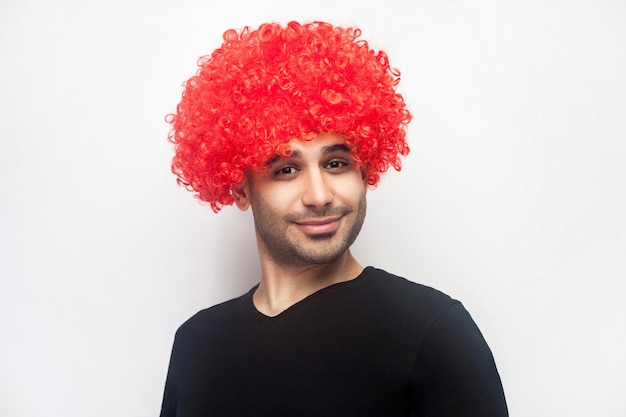 Portrait d'un homme hipster élégant et drôle avec des poils et une perruque rouge bouclée sur la tête souriant à la caméra à la recherche d'un mode de vie optimiste positif ludique tourné en studio intérieur isolé sur fond blanc