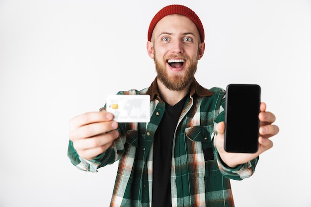 Portrait d'homme hipster tenant une carte de crédit et un téléphone portable, tout en se tenant isolé sur fond blanc