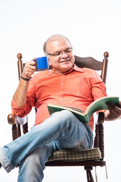 Portrait d'un homme indien ou asiatique mature heureux lisant un livre assis sur une chaise à bascule en sirotant du thé ou du café dans une tasse