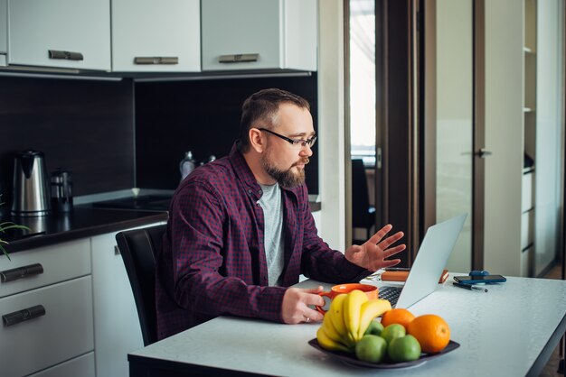 Portrait d'un homme intelligent élégant avec une barbe dans un intérieur de cuisine moderne. Un homme d'affaires en chemise travaille à la maison avec un ordinateur portable pendant la période de quarantaine. Communication avec des amis en isolement.