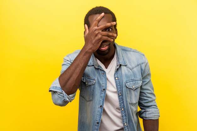 Portrait d'un homme intéressé positif en chemise décontractée en denim couvrant le visage et les yeux avec la main, regardant à travers les doigts avec une expression heureuse et curieuse. studio d'intérieur tourné isolé sur fond jaune