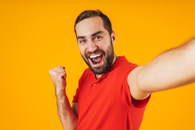 Portrait d'un homme joyeux en t-shirt rouge riant et se réjouissant tout en prenant une photo de selfie isolée sur jaune