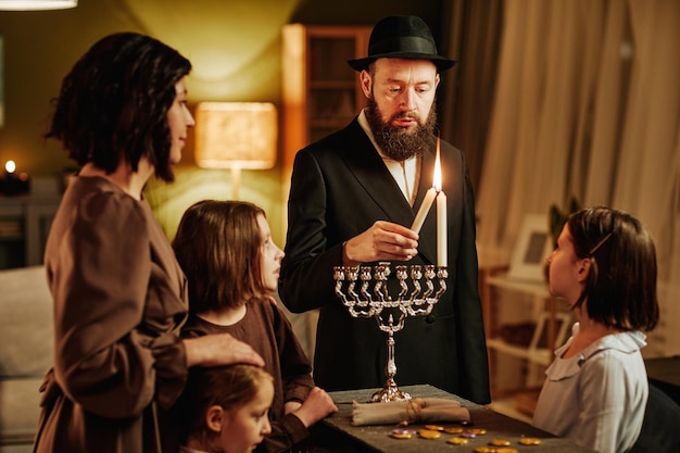 Portrait d'un homme juif orthodoxe avec une famille allumant une bougie menorah pendant la célébration de hanukkah