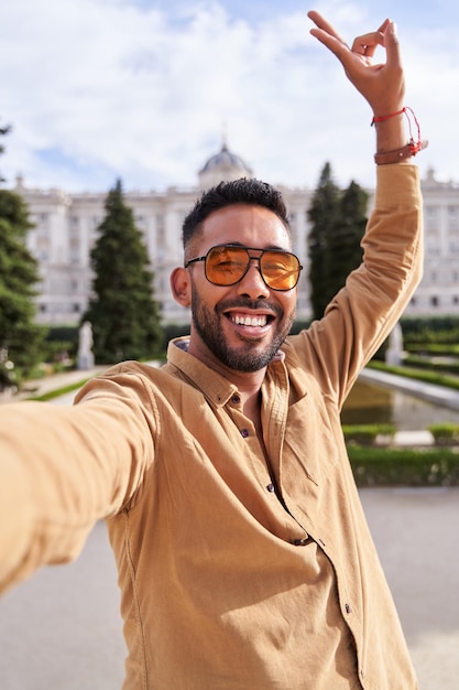 Photo portrait d'homme latin avec barbe et lunettes de soleil prenant un selfie avec son téléphone portable avec le palais royal de madrid espagne en arrière-plan