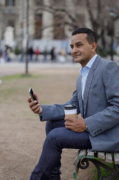 Portrait d'homme latino en veste avec une tasse de café jetable assis sur un banc dans un parc public