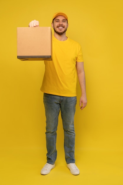 Portrait d'homme de livraison excité en uniforme jaune tenant une boîte de papier isolée sur mur jaune