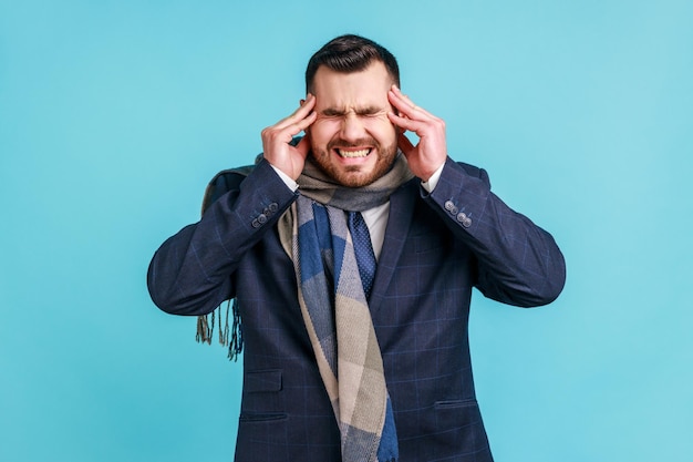 Portrait d'un homme malade barbu portant un costume de style officiel et enveloppé dans une écharpe souffrant de maux de tête et de migraine extrêmement forts. Studio intérieur tourné isolé sur fond bleu.