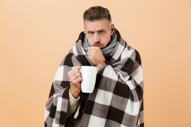 Portrait d'un homme malade enveloppé dans une couverture debout isolé sur mur beige, tenant une tasse avec du thé chaud