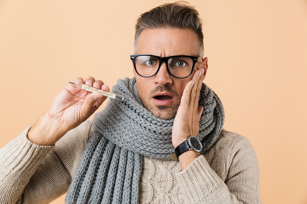 Portrait d'un homme malade habillé en pull et écharpe debout isolé sur mur beige, montrant un thermomètre