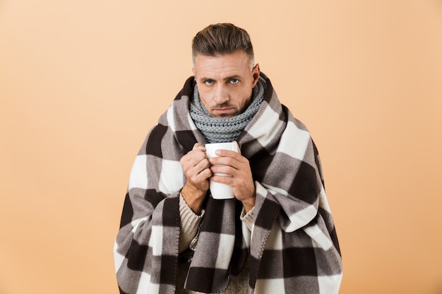 Portrait d'un homme malade triste enveloppé dans une couverture debout isolé sur mur beige, tenant une tasse avec du thé chaud