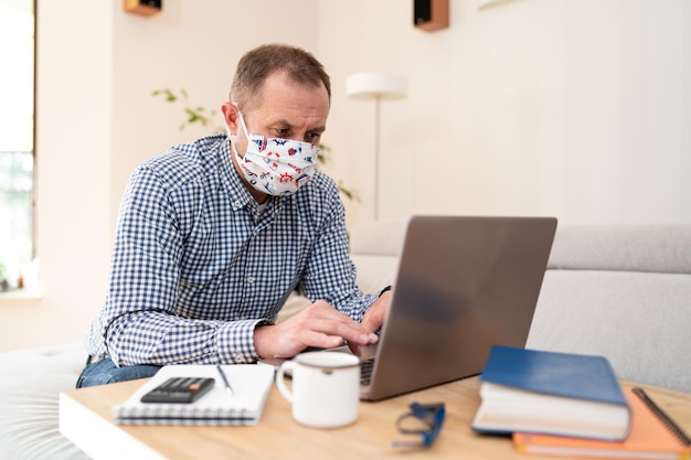 Portrait d'un homme avec un masque médical chirurgical assis et travaillant sur un ordinateur portable