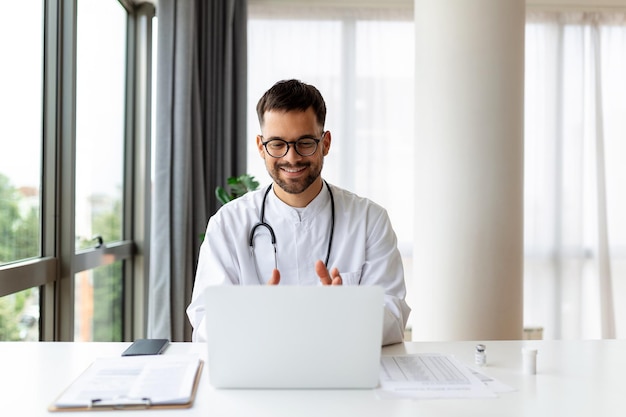Portrait d'un homme médecin parlant à un patient en ligne sur l'écran d'un ordinateur portable assis au bureau de la clinique donnant des consultations en ligne pour le traitement de santé domestique Télémédecine rendez-vous médical à distance