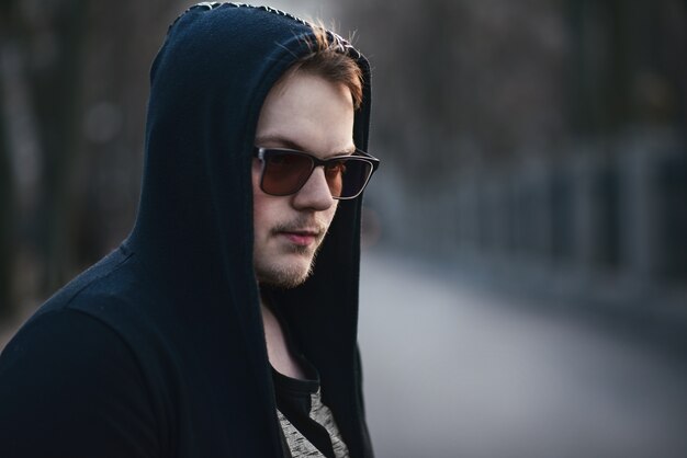 Portrait d'homme de mode dans un pull noir avec une capuche et des lunettes de soleil