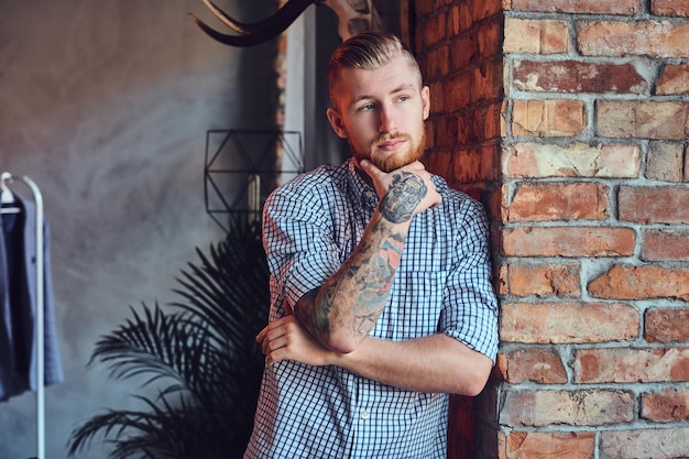 Portrait d'un homme moderne barbu avec des tatouages sur ses bras posant près de la fenêtre dans une pièce avec un intérieur loft.