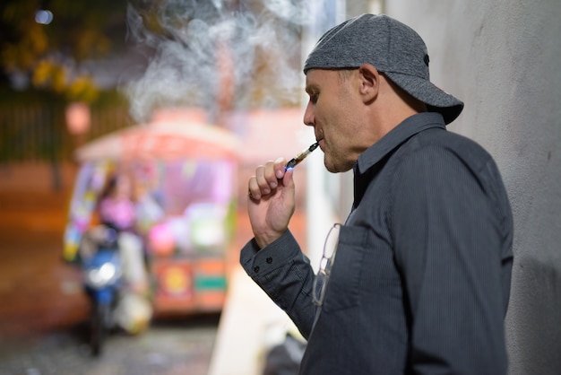 Portrait d'un homme mûr qui fume une cigarette électronique dans les rues la nuit