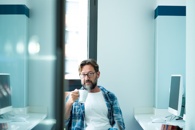 Portrait d'homme mûr regardant à la caméra tout en buvant du café au bureau - travail en ligne indépendant avec un style de vie de concept informatique - personnes de race blanche adultes dans un espace de coworking - couleurs d'humeur bleue personnes modernes