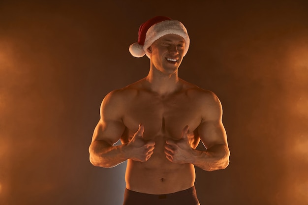 Portrait d'un homme musclé portant un bonnet de Noel de Noël, montrant le pouce vers le haut et le sourire sur fond enfumé