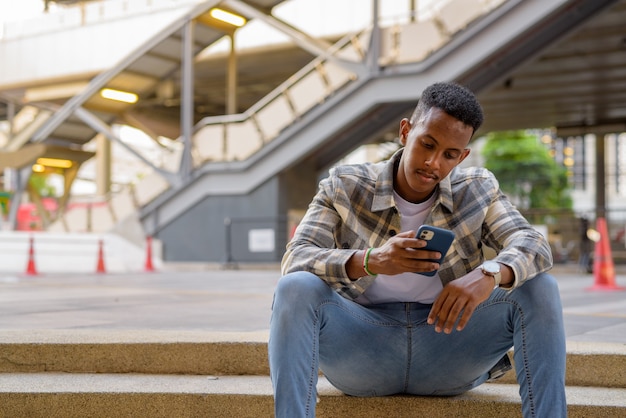 Portrait d'un homme noir africain assis à l'extérieur dans la ville pendant l'été à l'aide d'un plan horizontal de téléphone portable