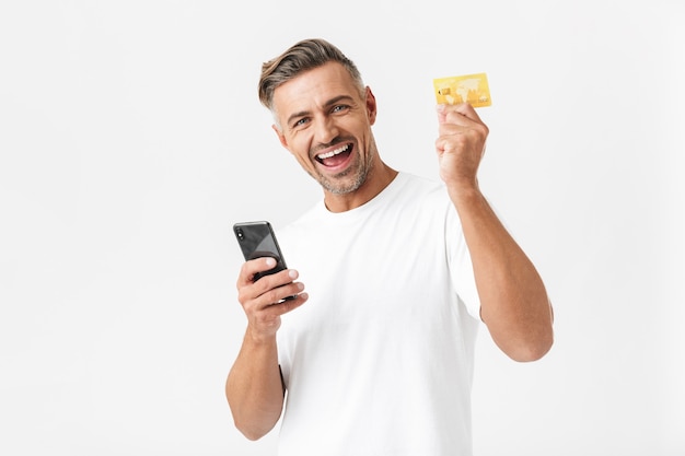 Portrait d'un homme optimiste des années 30 portant un t-shirt décontracté tenant un smartphone et une carte de crédit en plastique isolée sur blanc