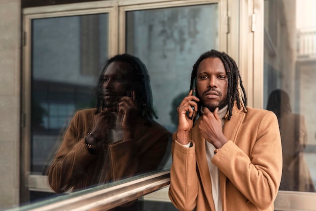 Portrait d'un homme pensif avec des dreadlocks modernes dans la ville parlant par téléphone portable reflet de son visage sur le mur de verre