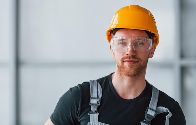 Portrait d'un homme portant des lunettes de protection uniformes grises et un casque orange qui se tient à l'intérieur dans un grand bureau moderne pendant la journée