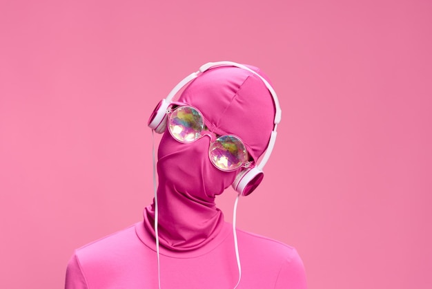 Portrait d'un homme portant un masque incognito rose avec des écouteurs et des lunettes de soleil rondes brillantes sur un fond rose Photographie d'art conceptuel Total rose Photo de haute qualité