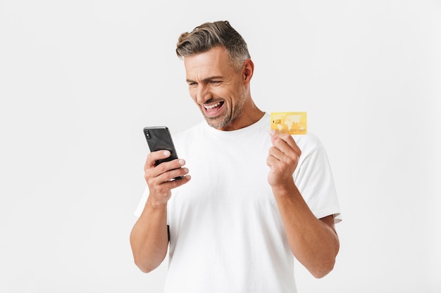 Portrait d'un homme positif des années 30 portant un t-shirt décontracté tenant un smartphone et une carte de crédit en plastique isolée sur blanc