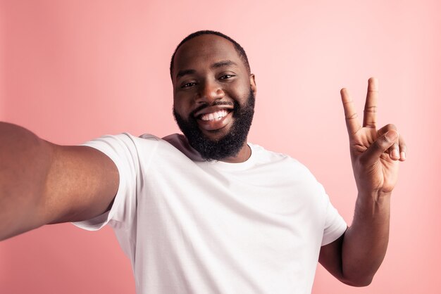 Portrait d'un homme positif gai prendre un selfie montrant un signe v sourire rayonnant sur un mur rose