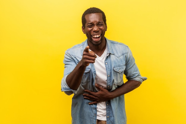 Portrait d'un homme positif joyeux en chemise en jean avec manches retroussées tenant la main sur le ventre et riant à haute voix pointant vers la caméra se moquant de vous tourné en studio intérieur isolé sur fond jaune