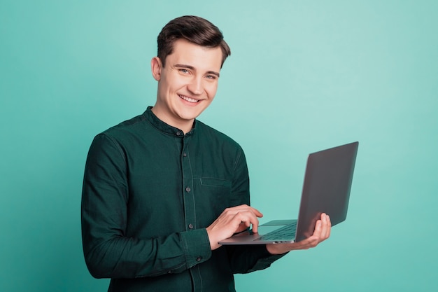 Portrait d'un homme programmeur charmant tenant un ordinateur portable en tapant un sourire à pleines dents sur fond bleu sarcelle