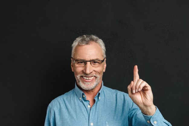 Portrait d'homme retraité caucasien des années 60 avec les cheveux gris et la barbe en chemise souriant et pointant l'index vers le haut sur copyspace, isolé sur mur noir