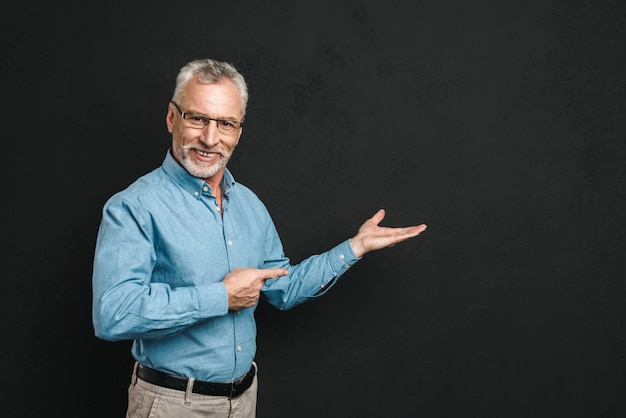 Portrait d'homme à la retraite heureux des années 60 avec les cheveux gris et la barbe en costume souriant et démontrant copyspace texte ou produit, isolé sur mur noir