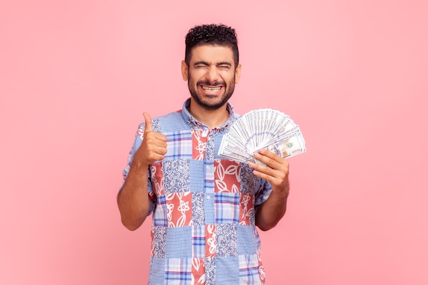 Portrait d'un homme satisfait avec barbe portant une chemise bleue de style décontracté tenant des billets en dollars et montrant les pouces vers le haut en profitant d'une vie riche Prise de vue en studio intérieure isolée sur fond rose