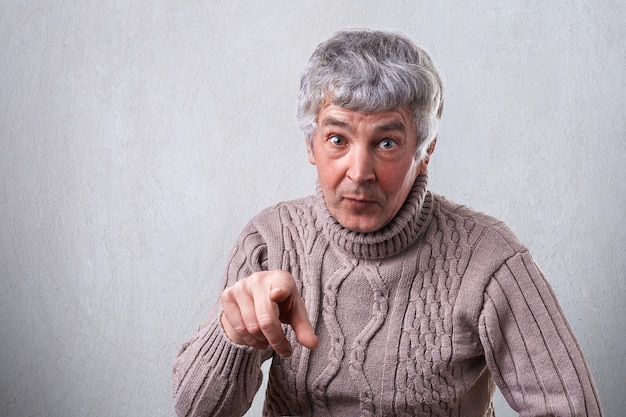 Un portrait d'un homme senior attrayant aux cheveux gris et aux rides ayant une expression surprise pointant vers vous avec son index