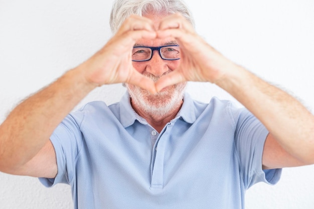 Portrait d'un homme senior aux cheveux blancs avec des lunettes regardant la caméra en forme de coeur avec les mains