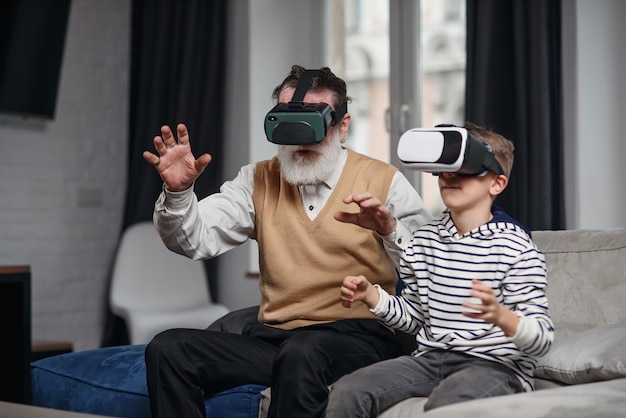 Portrait d'un homme senior excité à l'aide de lunettes VR assis sur un canapé à la maison avec un petit-fils qui rit à côté de lui