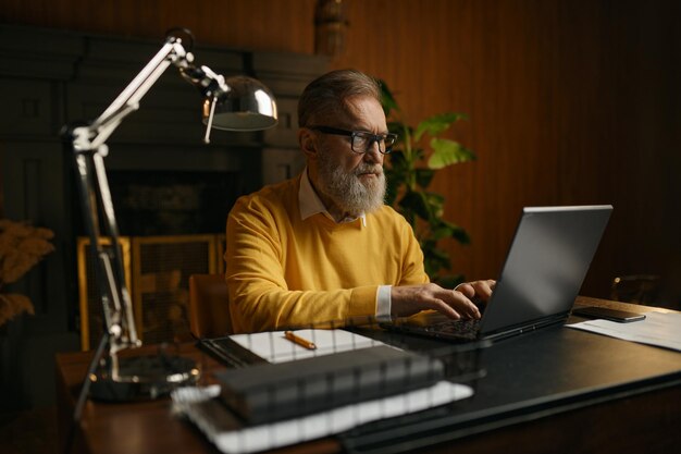 Portrait d'un homme senior tapant sur un clavier d'ordinateur portable assis à table et travaillant au bureau à domicile le soir