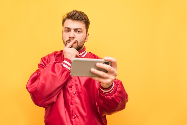 Portrait d'un homme sérieux avec une barbe, vêtu d'une veste rouge, se tient avec un smartphone à la main sur jaune