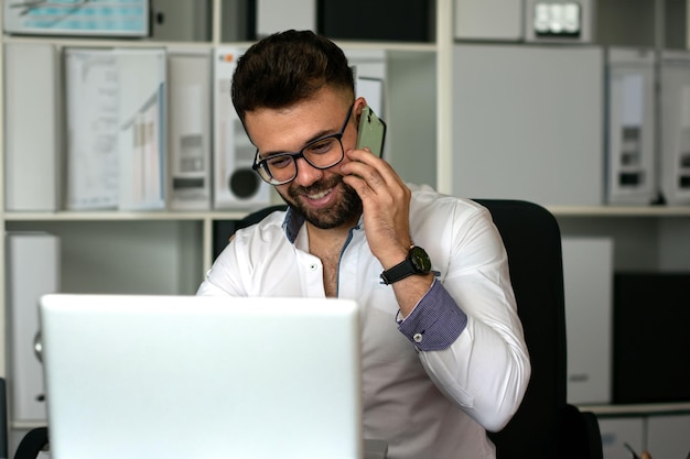 Portrait d'un homme souriant avec une barbe assis avec un ordinateur portable en train de parler au téléphone