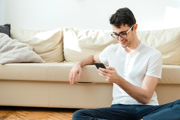 Portrait d'un homme souriant séduisant dans des vêtements décontractés assis sur le sol près du canapé dans le salon et regardant les nouvelles à l'aide d'un téléphone portable