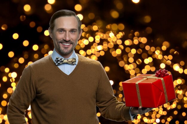 Portrait d'un homme souriant tenant une boîte cadeau rouge. Charmant homme caucasien mature et positif avec un cadeau de Noël sur fond avec des lumières rougeoyantes.