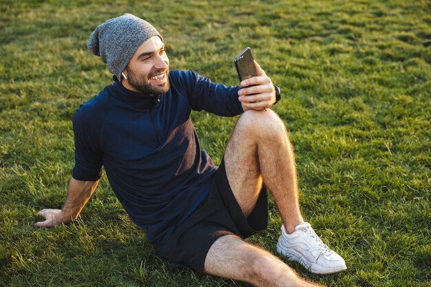 Portrait d'un homme sportif souriant vêtu d'un survêtement utilisant un smartphone et assis sur l'herbe pendant l'entraînement dans un parc verdoyant